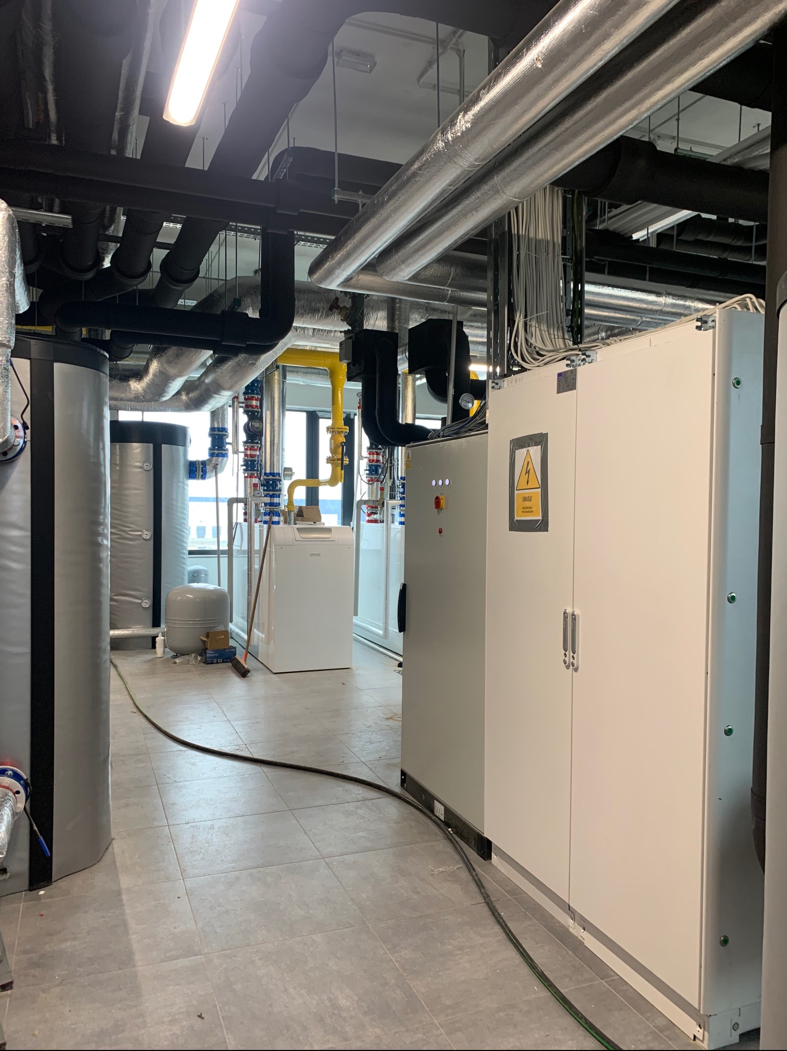 Węzeł cieplny z kotłem gazowym, zbiornikami i szafą sterowniczą. Rurociągi pod stropem, instalacje w kolorach: czarnym, srebrnym i żółtym. Widok na okno.