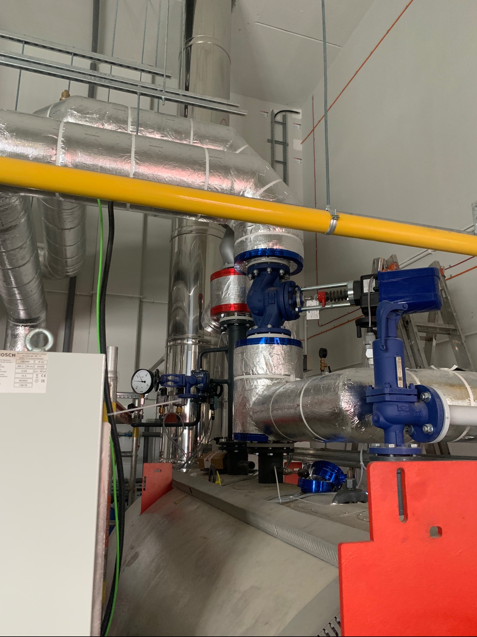 Złożona instalacja przemysłowa z rurami w srebrnej izolacji, niebieskimi zaworami i żółtą rurą. Widoczny manometr i drabina w tle. Elementy instalacji częściowo poza kadrem.