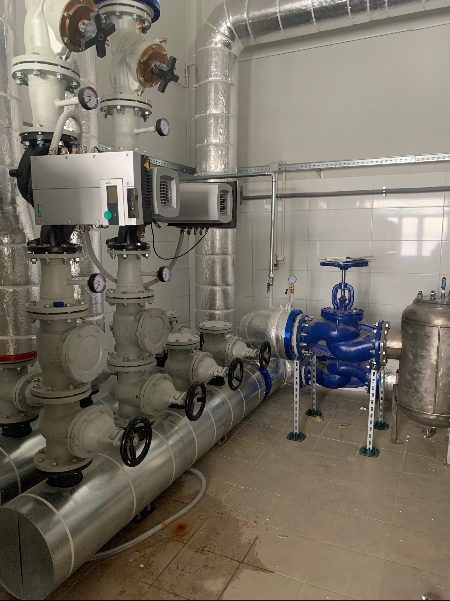 Instalacja przemysłowa z rurami i zaworami w Środzie Wielkopolskiej. Rury w izolacji, manometry, białe płytki na ścianie. Elementy instalacji w kolorze niebieskim.