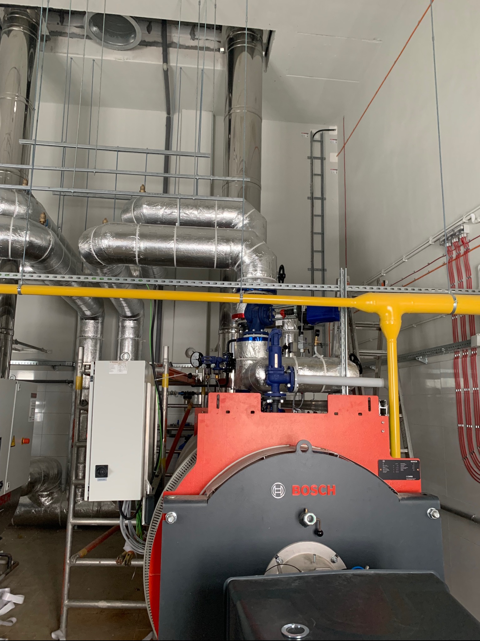 Nowoczesna kotłownia z piecem Bosch, rurami w izolacji i żółtą instalacją gazową. Widoczne przewody elektryczne i drabina. Białe ściany i sufit.
