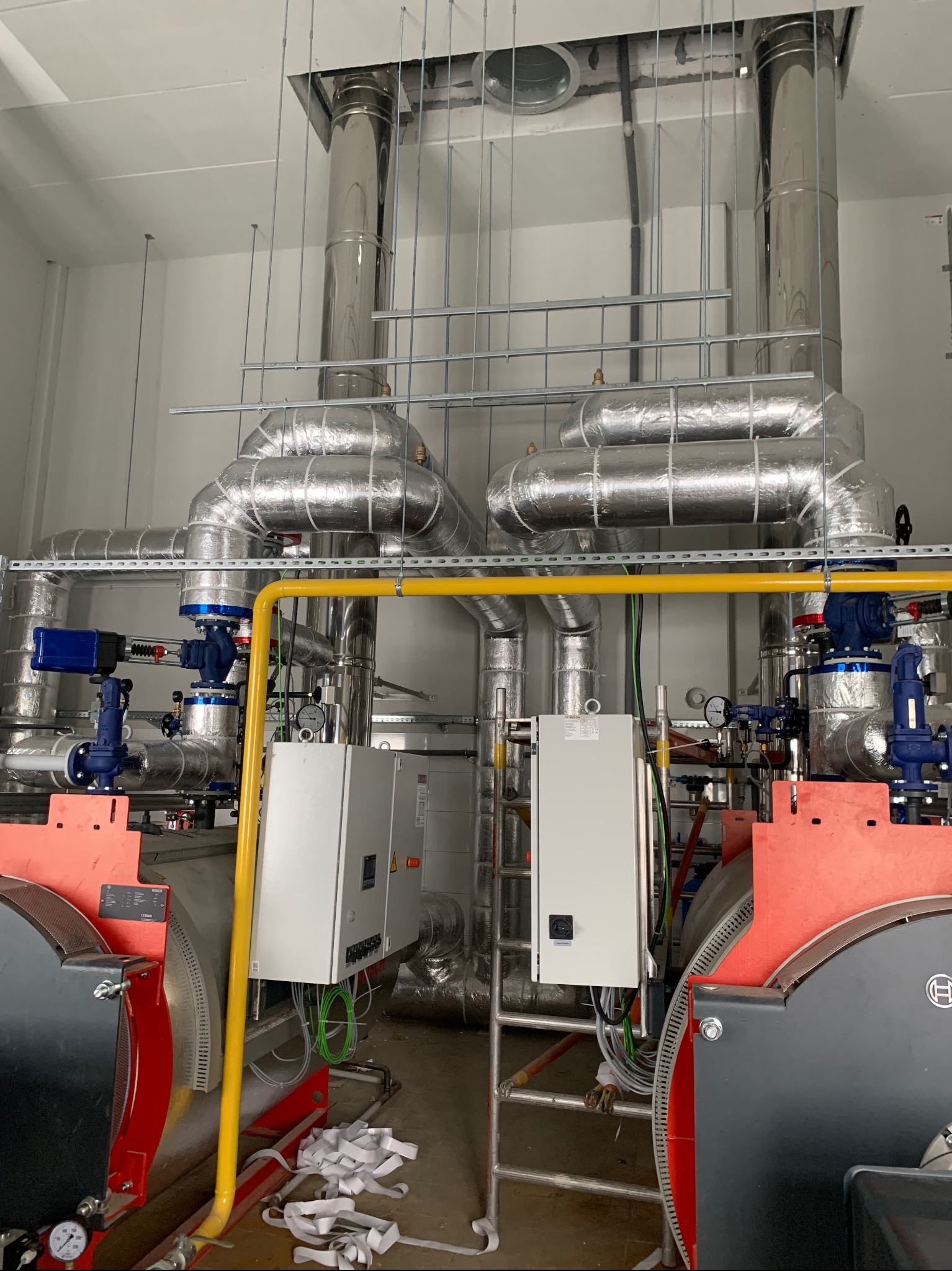 Dwa kotły grzewcze Bosch z izolowanymi rurami, zaworami i żółtą instalacją gazową w jasnym pomieszczeniu technicznym. Widoczne elementy sterujące i drabina.
