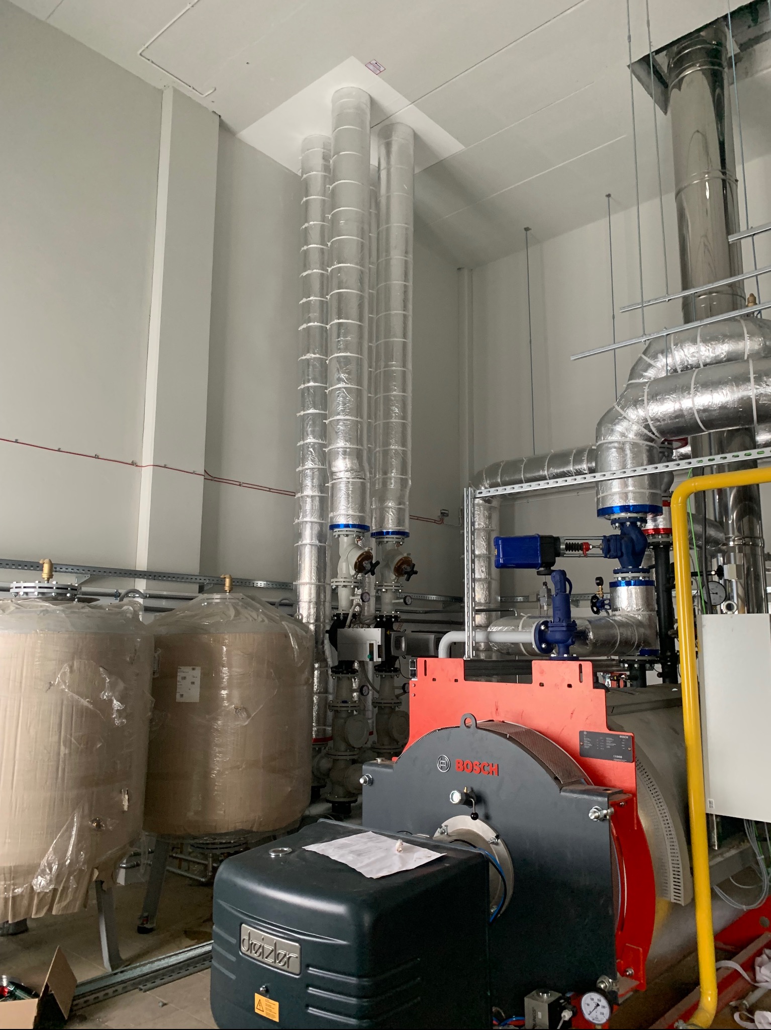 Nowoczesna kotłownia z piecem Bosch, zbiornikami i izolowanymi rurami. Widoczne elementy instalacji w srebrnej izolacji i rury odprowadzające spaliny.