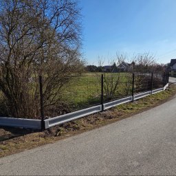 Redshop Wojciech Ustrzycki - Nowoczesne ogrodzenie panelowe z podmurówką w Krakowie. Ciemna siatka na betonowej podstawie, widok z drogi, błękitne niebo w tle. Solidne i estetyczne.