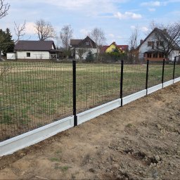 Redshop Wojciech Ustrzycki - Nowoczesne ogrodzenie panelowe z podmurówką betonową na nierównym terenie. Czarne słupki i siatka kontrastują z jasną podmurówką. Widok na domy w tle.