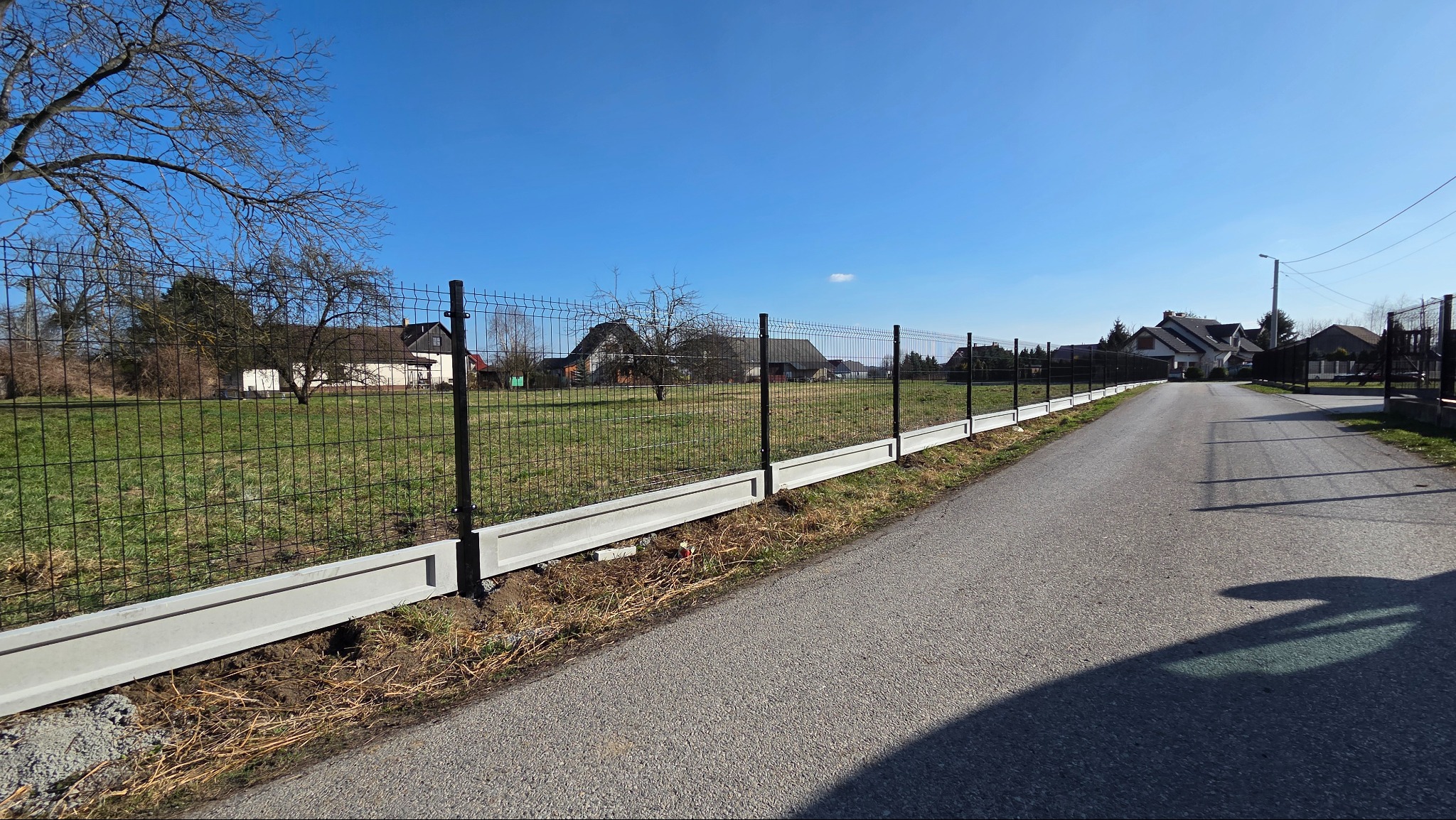 Nowoczesne ogrodzenie panelowe z podmurówką betonową, widok z perspektywy ulicy, słoneczny dzień, błękitne niebo, domy w tle. Minimalistyczny design.