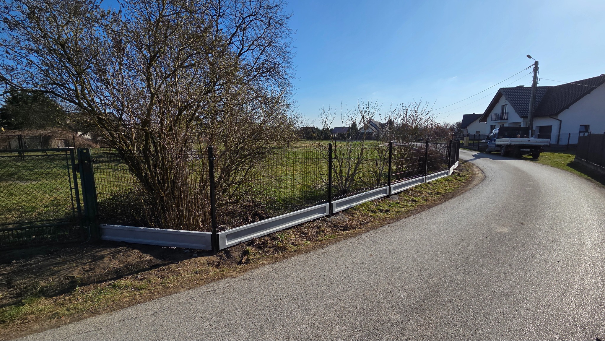 Nowoczesne ogrodzenie panelowe z podmurówką w Krakowie. Ciemna siatka na betonowej podstawie, widok z drogi, błękitne niebo w tle. Solidne i estetyczne.