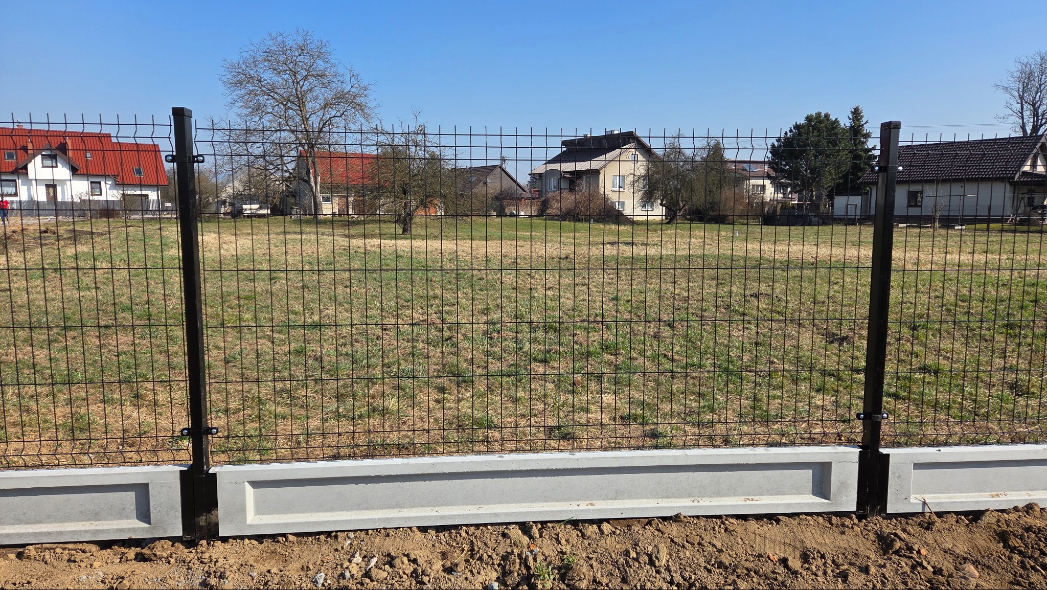 Nowoczesne ogrodzenie panelowe z podmurówką betonową na tle domów jednorodzinnych i błękitnego nieba. Widok z bliska na fragment ogrodzenia.