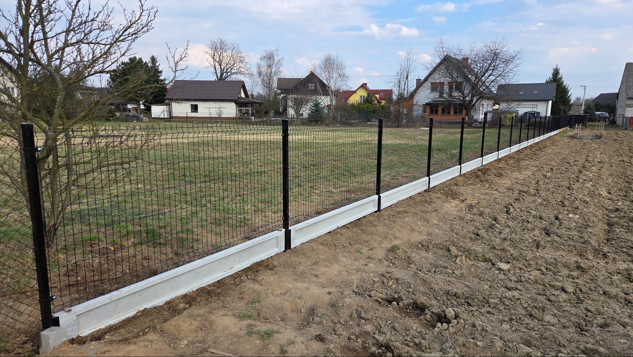 Nowoczesne ogrodzenie panelowe z podmurówką betonową na nierównym terenie. Czarne słupki i siatka kontrastują z jasną podmurówką. Widok na domy w tle.