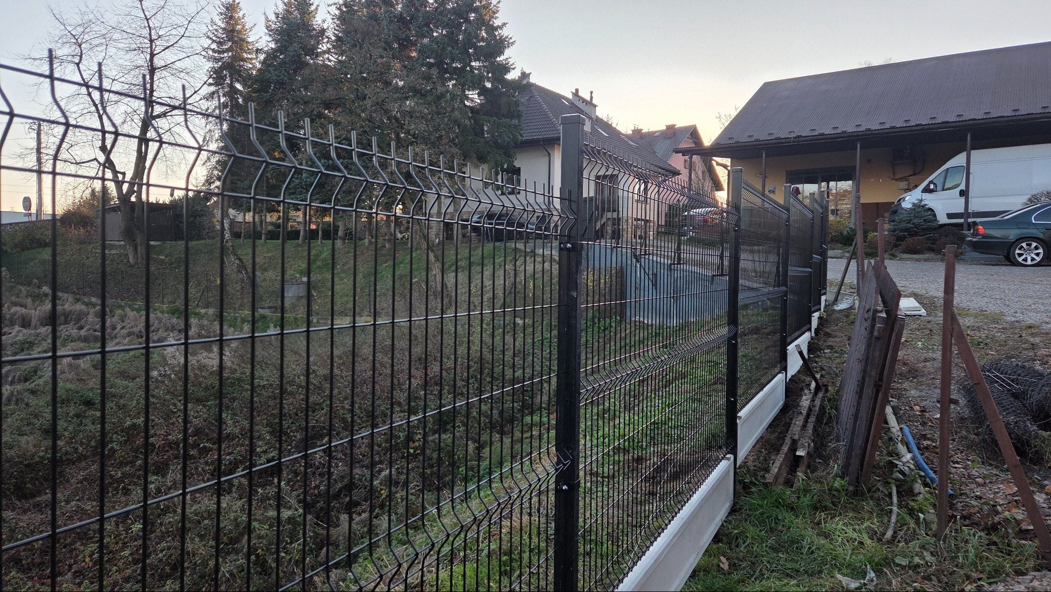 Czarny panel ogrodzeniowy na nierównym terenie, z widocznymi elementami montażowymi i podmurówką. W tle budynki mieszkalne i zieleń.