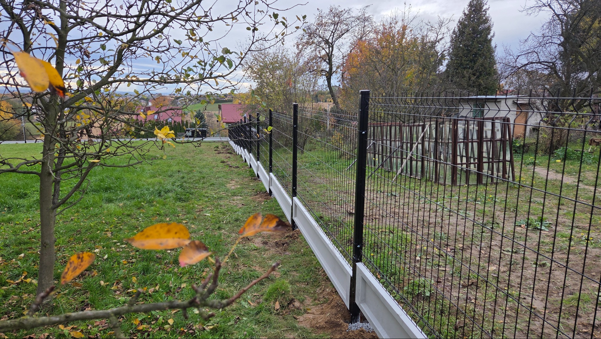 Nowoczesne ogrodzenie panelowe z podmurówką betonową na nierównym terenie, widok z bliska na fragment ogrodzenia z drzewem w tle. Jesienne liście na pierwszym planie.