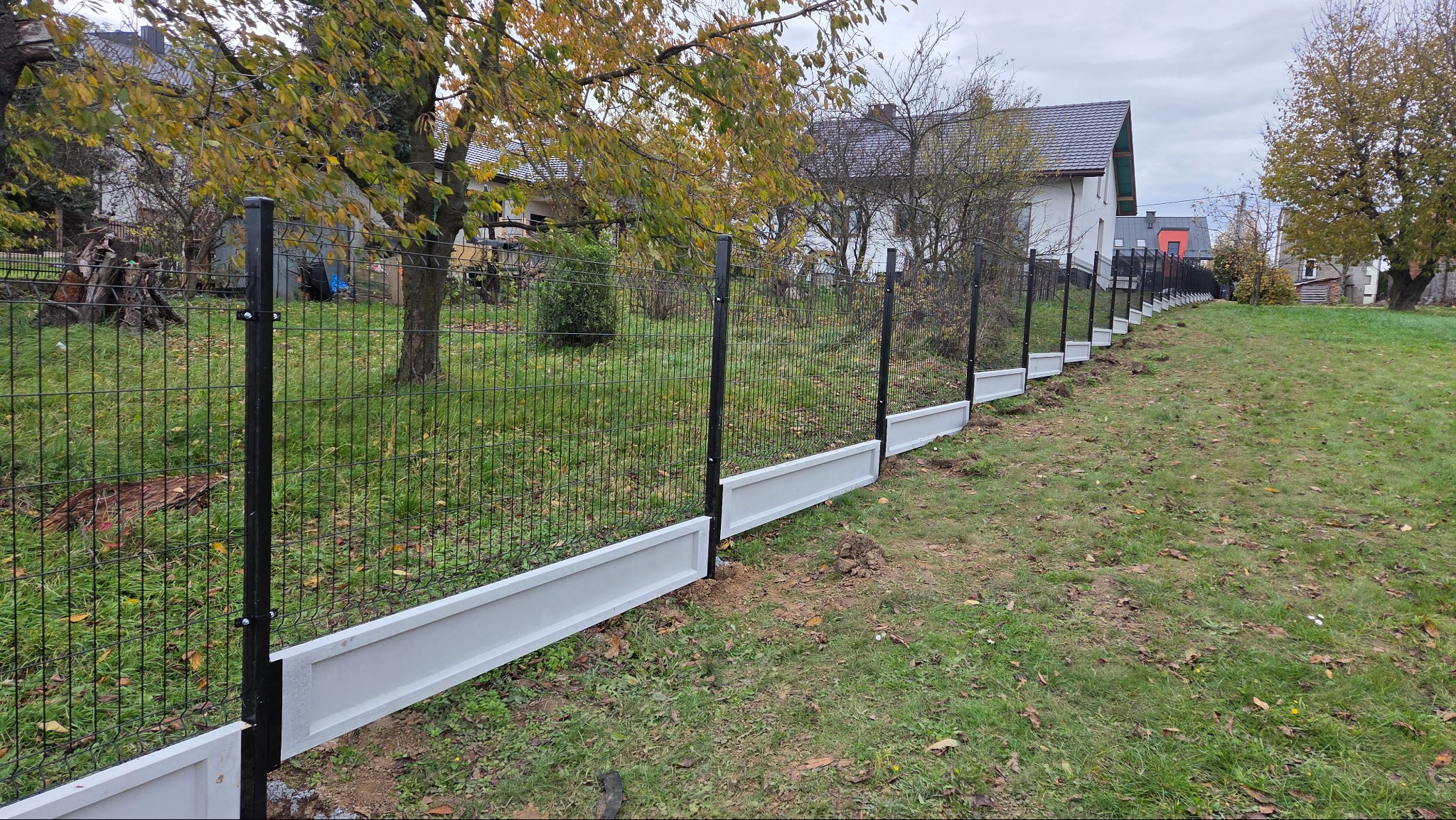 Nowoczesne ogrodzenie panelowe z podmurówką w kolorze białym, czarne słupki, widok na posesję z zielonym trawnikiem i drzewami w tle.