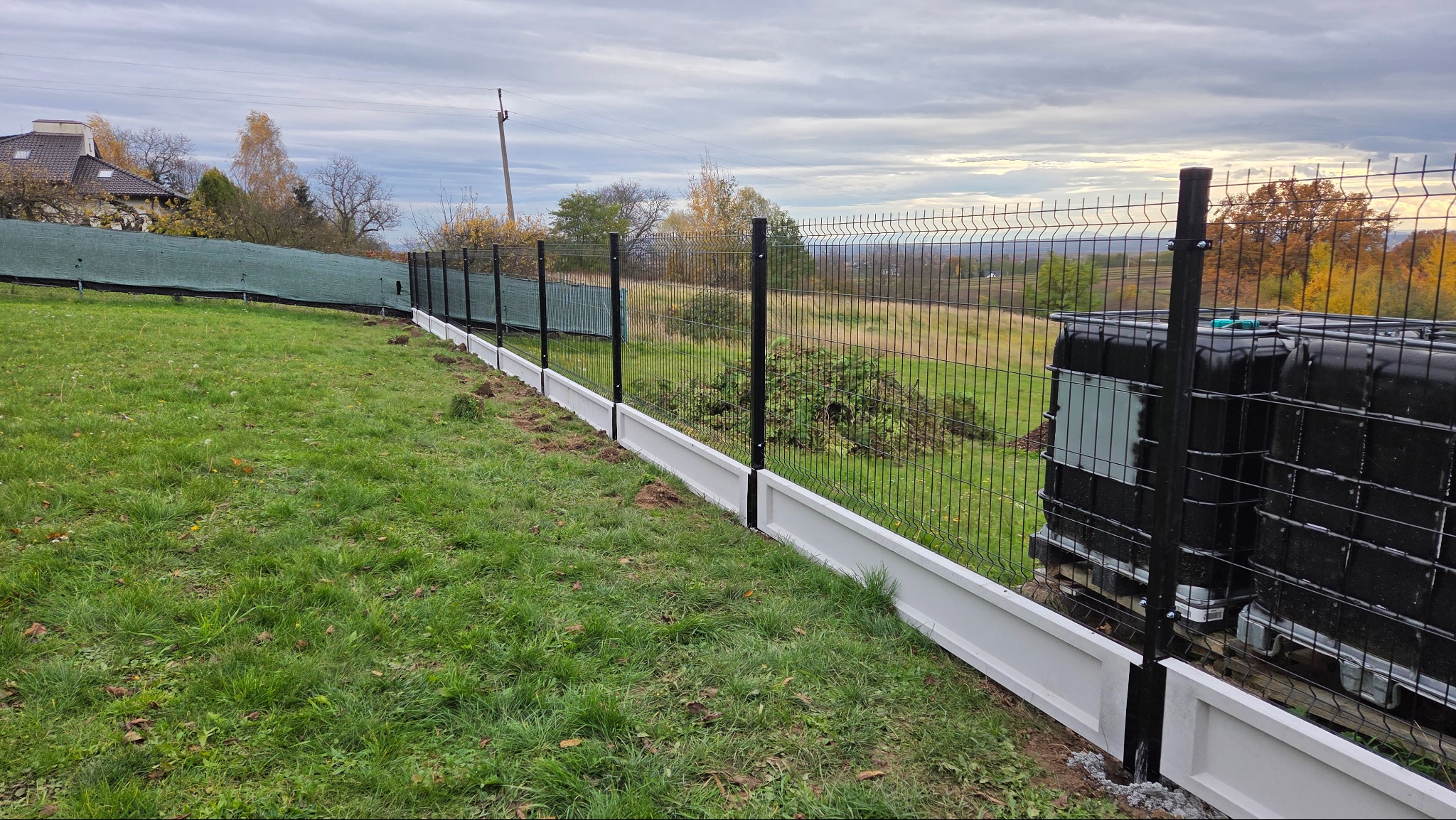 Nowoczesne ogrodzenie panelowe z podmurówką w kolorze białym, na tle zielonego trawnika i jesiennego krajobrazu. Widoczne zbiorniki na wodę za ogrodzeniem.
