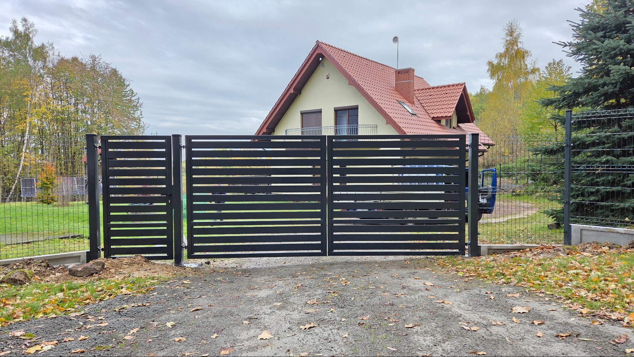 Nowoczesna, pozioma brama wjazdowa z czarnych paneli na tle domu z czerwoną dachówką i zieleni. Widoczna siatka ogrodzeniowa i jesienne drzewa.