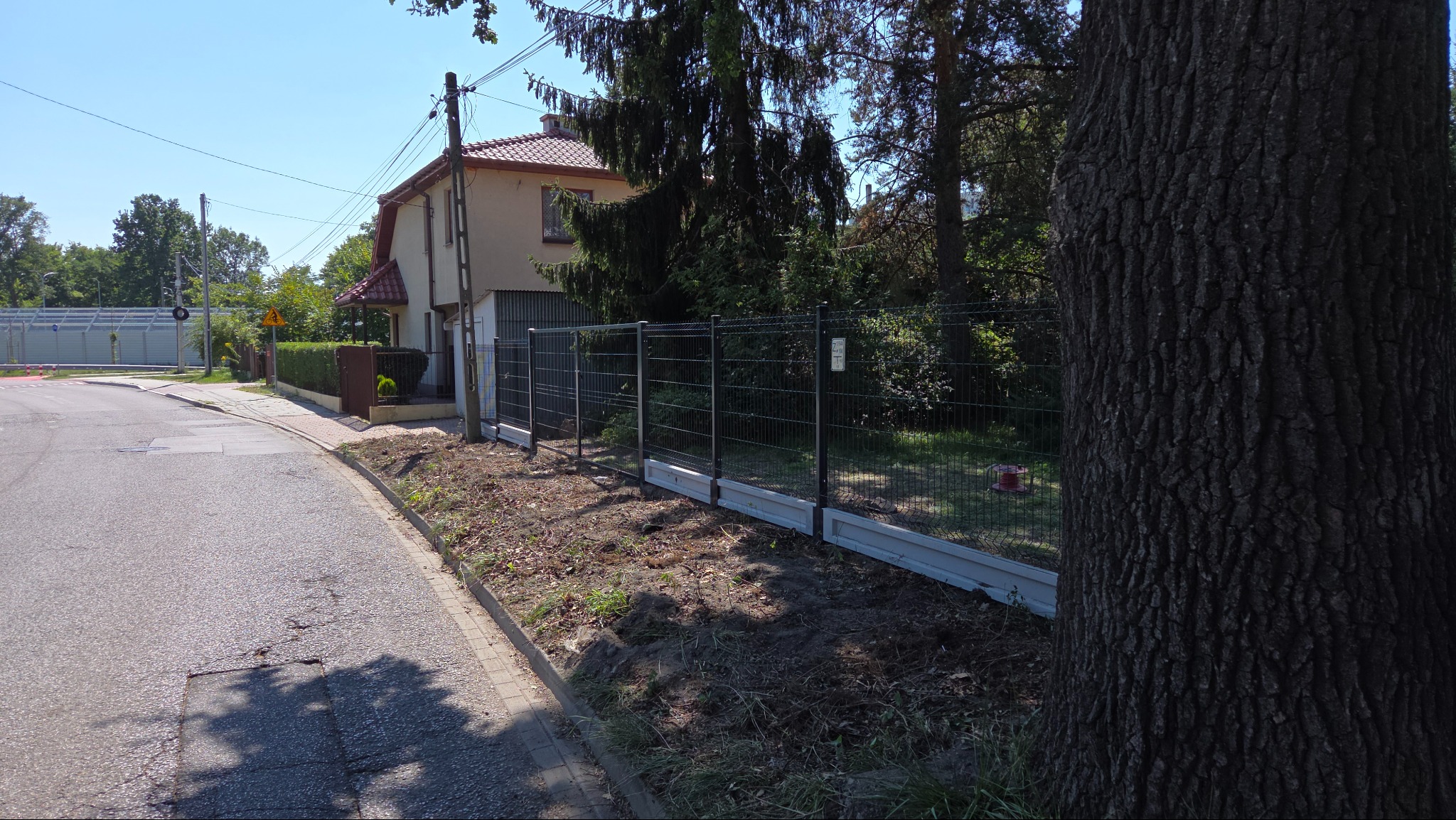 Nowoczesne ogrodzenie panelowe z podmurówką, widoczne z perspektywy ulicy, z domem w tle. Elementy ogrodzenia w kolorze grafitowym, podmurówka szara.