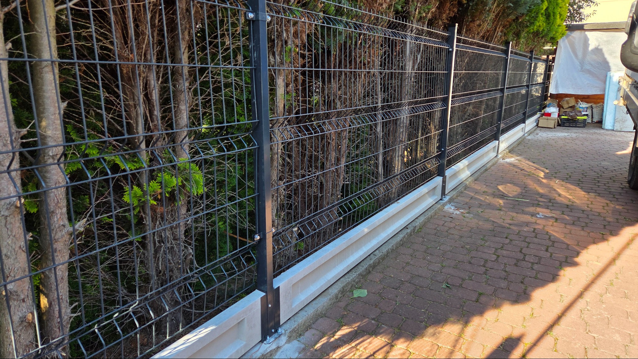 Nowoczesne ogrodzenie panelowe z podmurówką betonową, widoczne z perspektywy chodnika z kostki brukowej. Tło stanowią drzewa i krzewy.