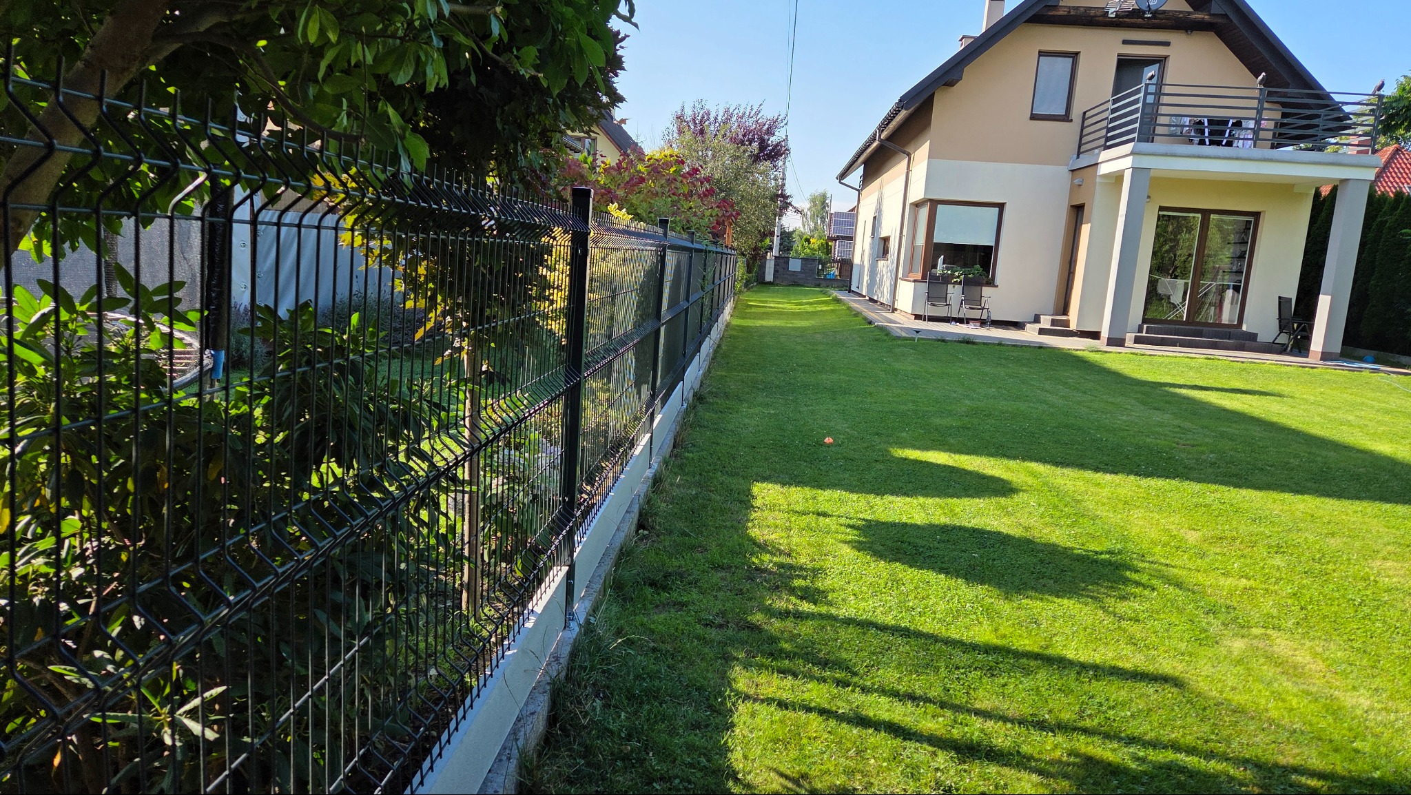 Nowoczesne ogrodzenie panelowe w kolorze grafitowym, oddzielające zadbany trawnik od posesji. Widok z perspektywy ogrodu, w tle dom z balkonem.