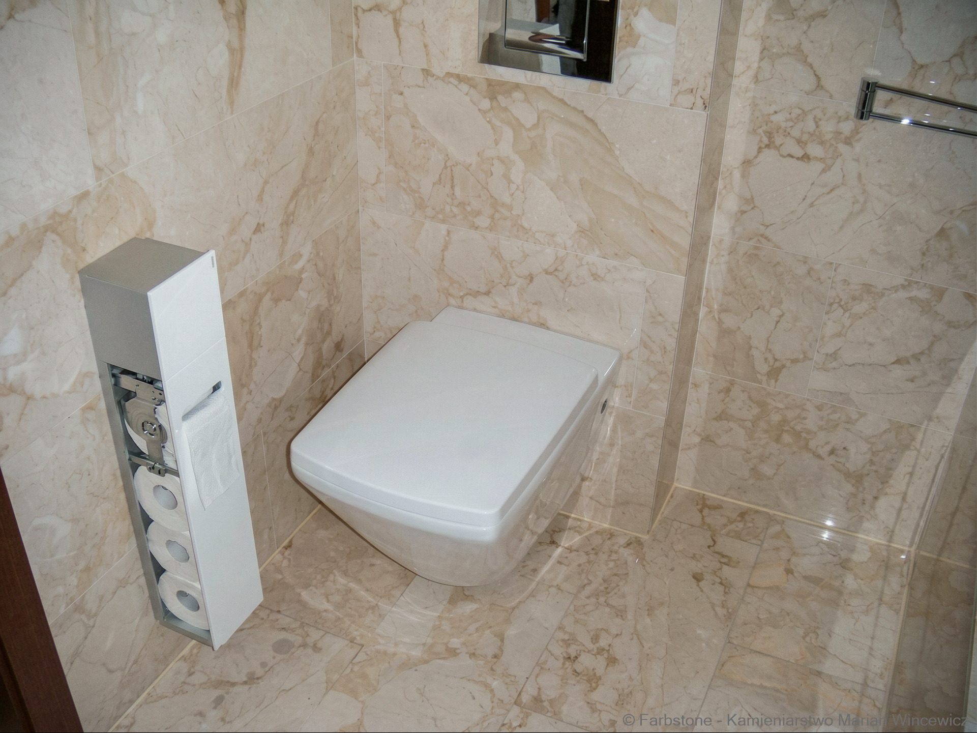 Beżowe płytki imitujące marmur w łazience w Warszawie. Wisząca toaleta i pojemnik na papier toaletowy. Widoczne fugi i chromowane elementy.