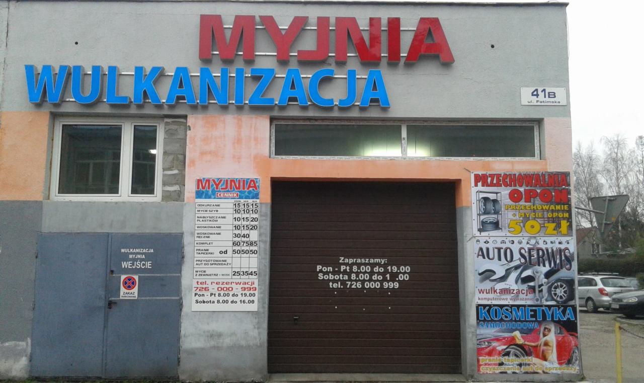 Fasada budynku z szyldami 'Myjnia' i 'Wulkanizacja', cennikiem usług, informacją o godzinach otwarcia i reklamą przechowalni opon.