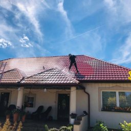 Przedsiębiorstwo Wielobranżowe Mateusz Orłowski - Pracownik w czarnym kombinezonie czyści czerwoną dachówkę domu jednorodzinnego za pomocą myjki ciśnieniowej. Widoczne smugi wody i błękitne niebo.