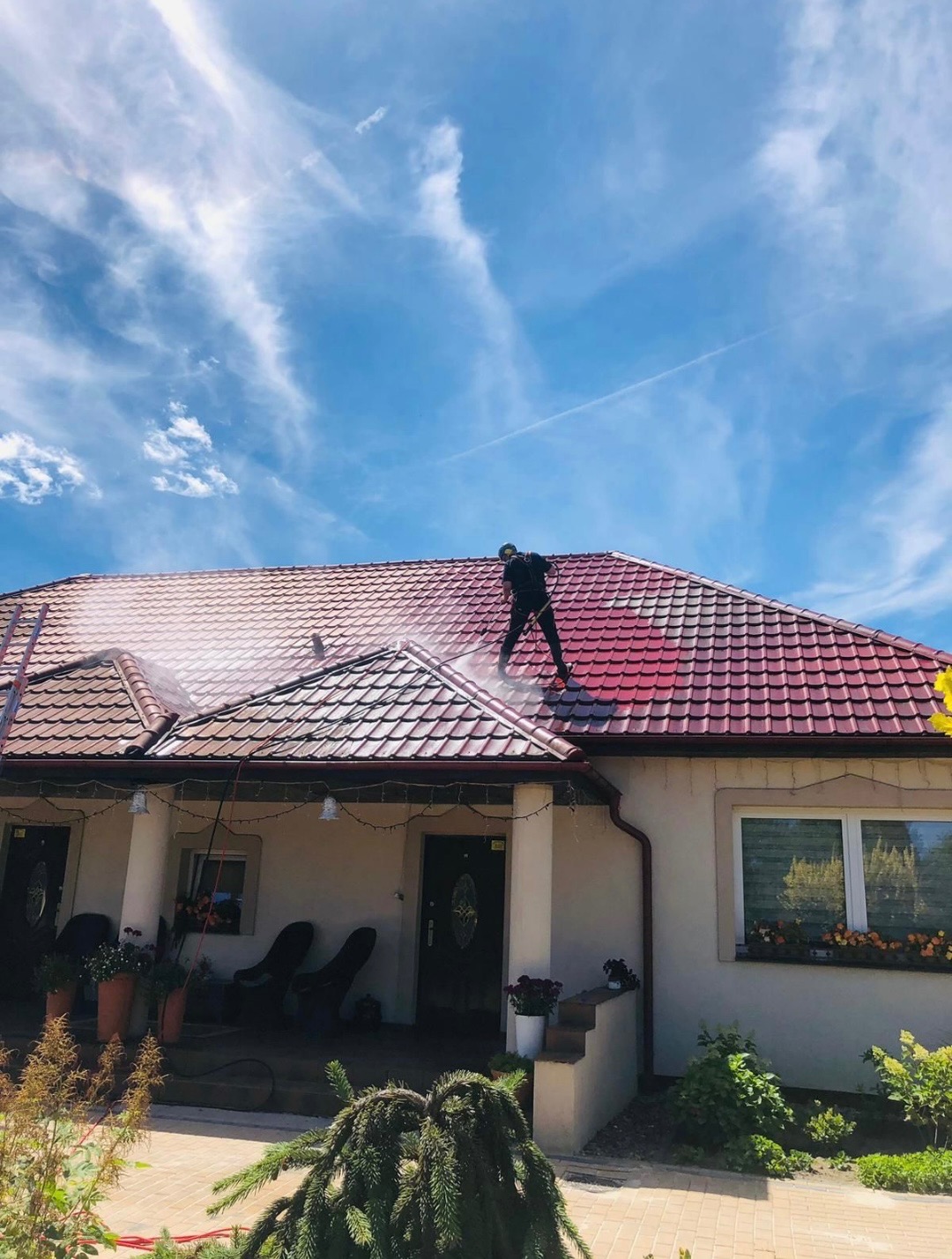 Pracownik w czarnym kombinezonie czyści czerwoną dachówkę domu jednorodzinnego za pomocą myjki ciśnieniowej. Widoczne smugi wody i błękitne niebo.