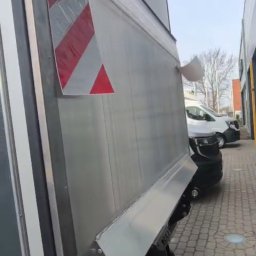 Dmytro Sveleba - Srebrny bus dostawczy z oznaczeniem ostrzegawczym na boku, zaparkowany na brukowanym placu, z widocznym fragmentem innego pojazdu w tle.