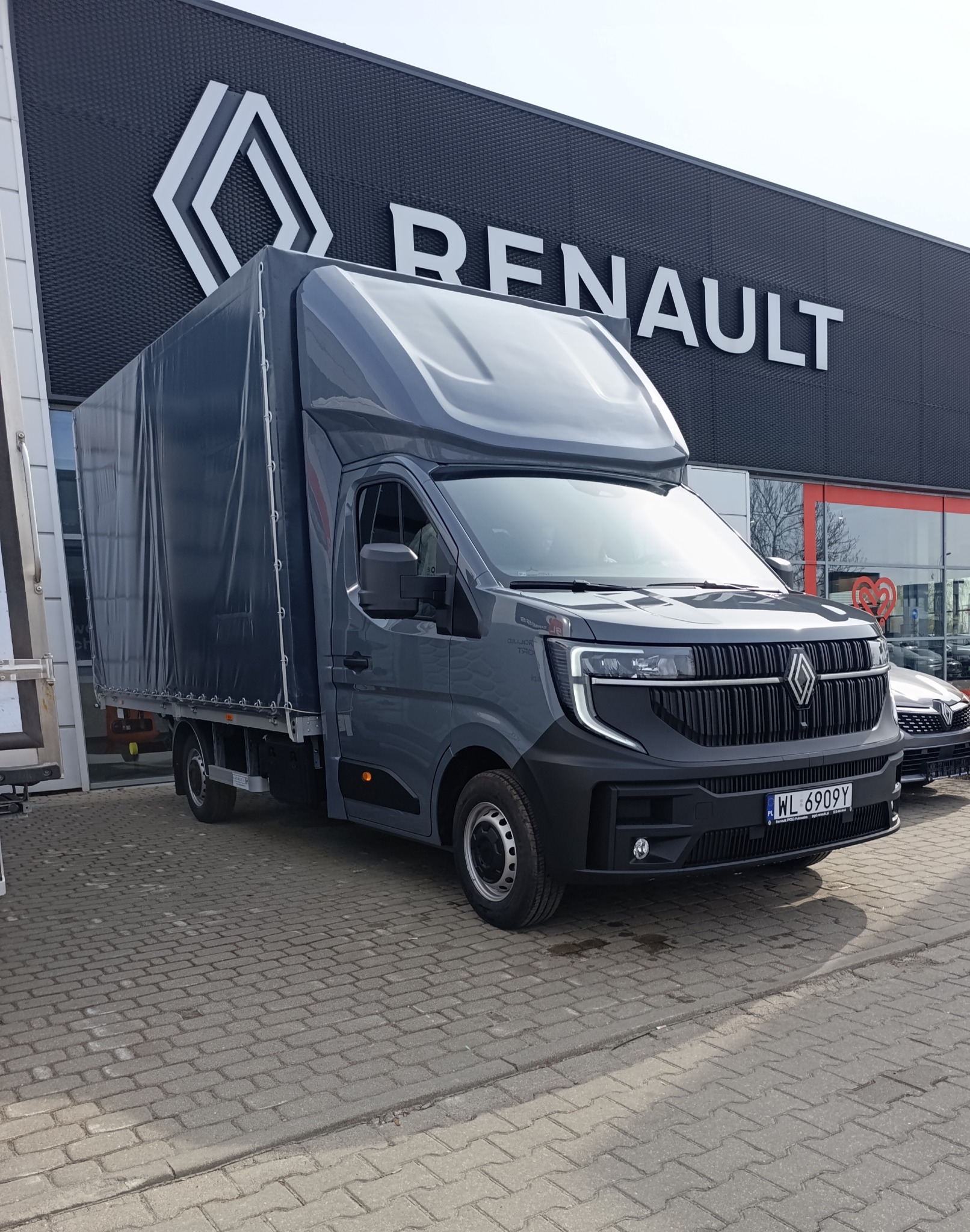 Szary bus Renault z plandeką na tle salonu samochodowego. Widoczny numer rejestracyjny. Prezentacja pojazdu gotowego do transportu.