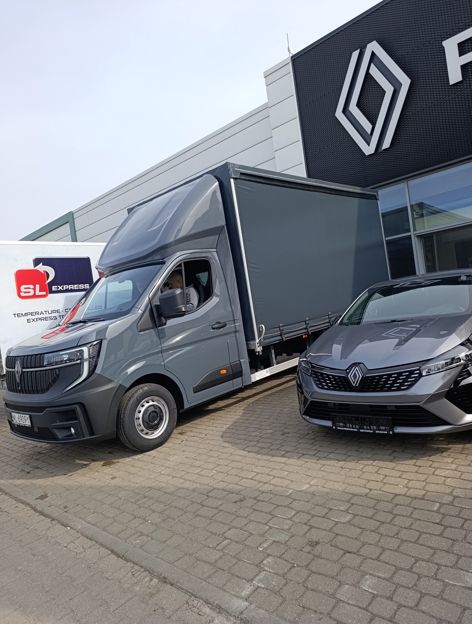 Szary bus dostawczy z plandeką zaparkowany obok szarego samochodu osobowego na tle salonu Renault. Widoczny kierowca w kabinie.