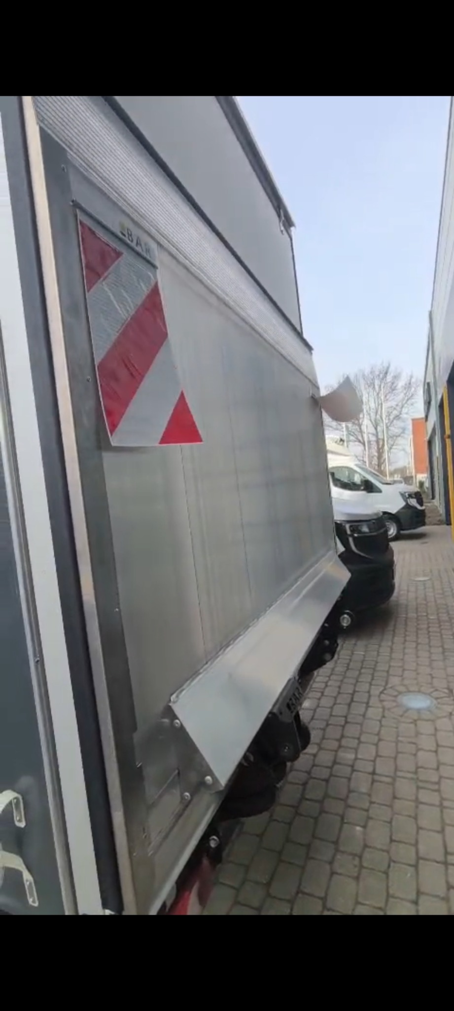 Srebrny bus dostawczy z oznaczeniem ostrzegawczym na boku, zaparkowany na brukowanym placu, z widocznym fragmentem innego pojazdu w tle.