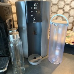 Mateusz Bajerski - Nowoczesny dystrybutor wody z wyświetlaczem temperatury, obok szklana butelka i pojemnik na wodę. Marmurowe płytki w tle.