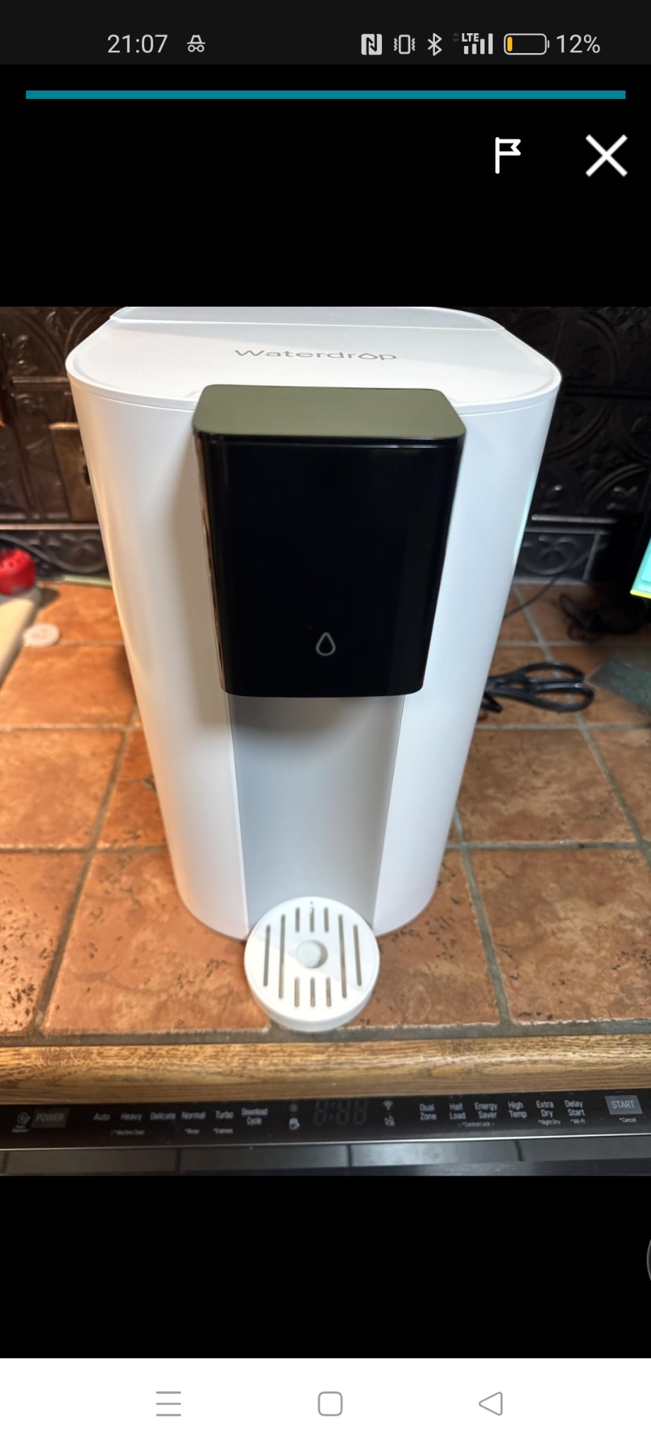 Dystrybutor wody Waterdrop w białej obudowie z czarnym panelem, umieszczony na blacie z brązowych płytek. Widoczny fragment zmywarki.