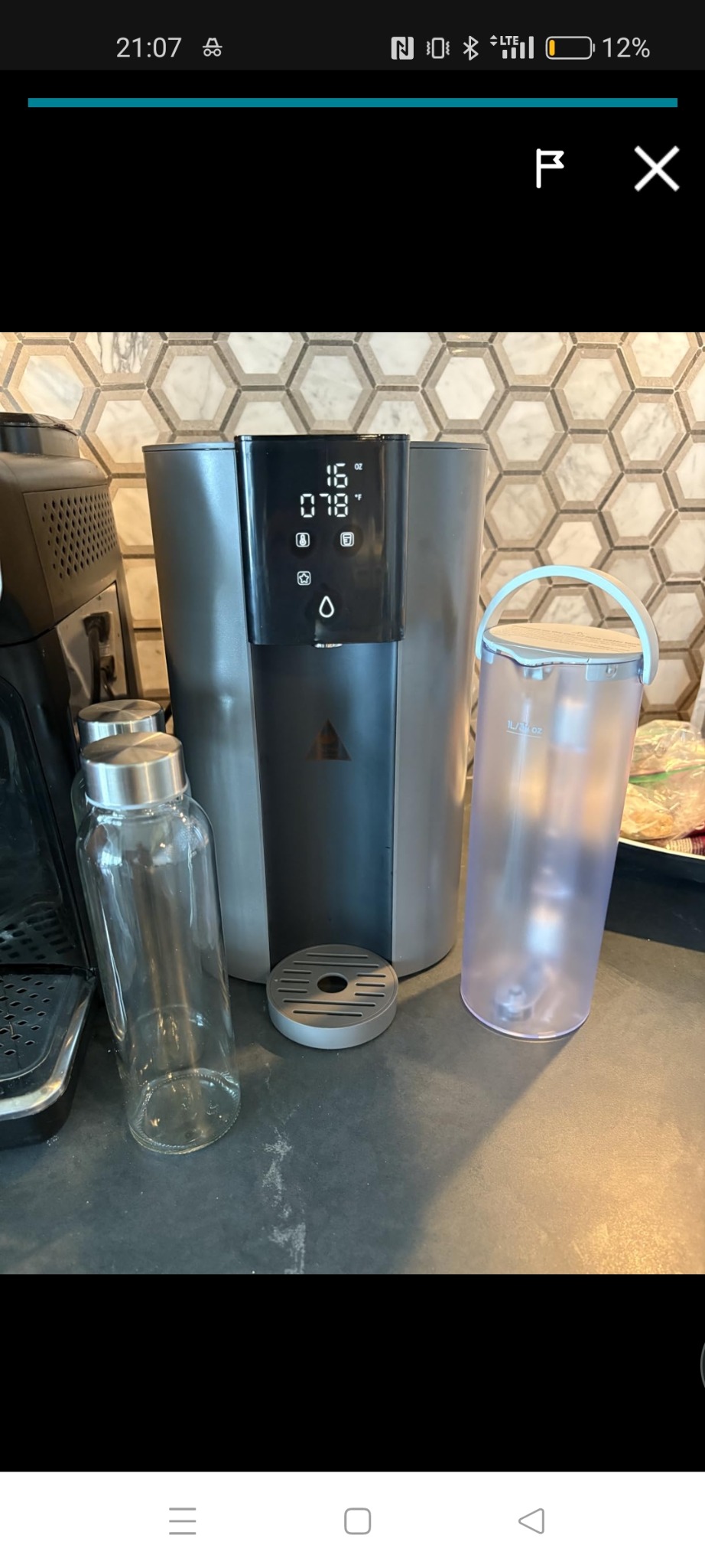 Nowoczesny dystrybutor wody z wyświetlaczem temperatury, obok szklana butelka i pojemnik na wodę. Marmurowe płytki w tle.