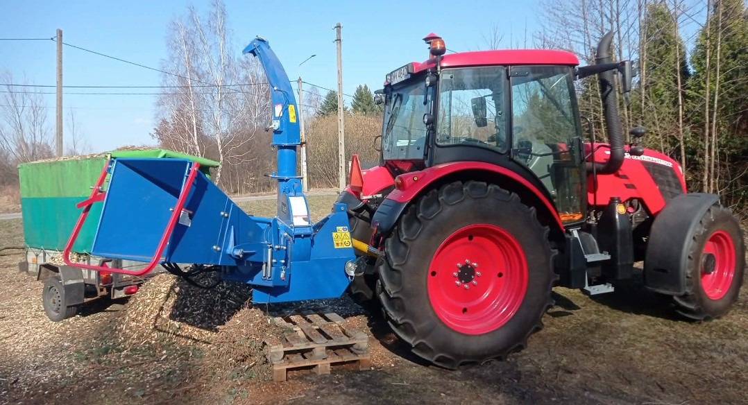Czerwony traktor z niebieskim rębakiem do gałęzi i przyczepą wypełnioną zrębkami na tle drzew. Sprzęt do rozdrabniania gałęzi po wycince.