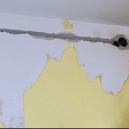 Volodymyr Hrytsyshyn - Bruzdowany przewód elektryczny w ścianie z widocznym gniazdkiem i puszką instalacyjną. Ściana w trakcie remontu, z resztkami farby i gładzi.