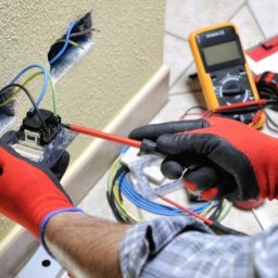 Volodymyr Hrytsyshyn - Elektryk w rękawicach montuje gniazdko elektryczne w ścianie. Widoczne przewody, miernik, notatnik i narzędzia. Ujęcie z bliska, skupione na dłoniach i instalacji.