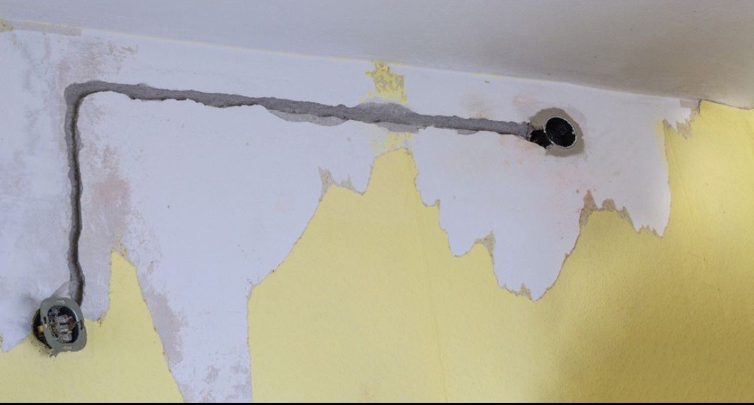 Bruzdowany przewód elektryczny w ścianie z widocznym gniazdkiem i puszką instalacyjną. Ściana w trakcie remontu, z resztkami farby i gładzi.