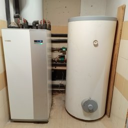 BILBO SPÓŁKA Z OGRANICZONĄ ODPOWIEDZIALNOŚCIĄ - Biała pompa ciepła Nibe i zbiornik akumulacyjny w kotłowni z beżowymi płytkami. Widoczne rury i zawory. Ujęcie z bliska, prezentujące instalację.