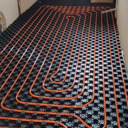 BILBO SPÓŁKA Z OGRANICZONĄ ODPOWIEDZIALNOŚCIĄ - Pomarańczowe rury ogrzewania podłogowego ułożone na czarnej macie w surowym wnętrzu. Widoczne elementy instalacji przy ścianie. Ujęcie z góry, szeroki kadr.