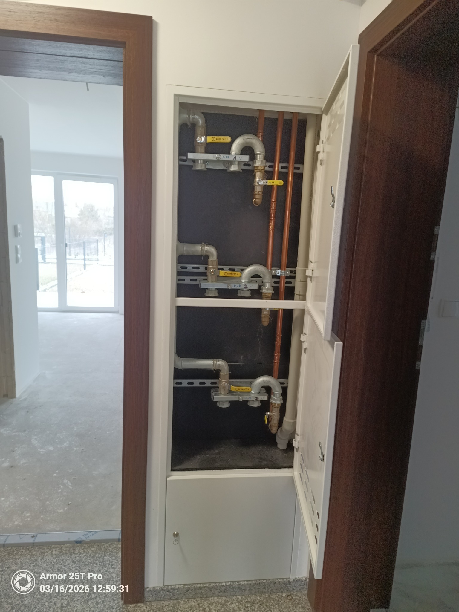 Otwarta szafka z pionowymi rurami miedzianymi i szarymi rurami instalacji gazowej, zamontowanymi na szynach. Widoczne żółte zawory. W tle surowy stan wykończenia wnętrza.