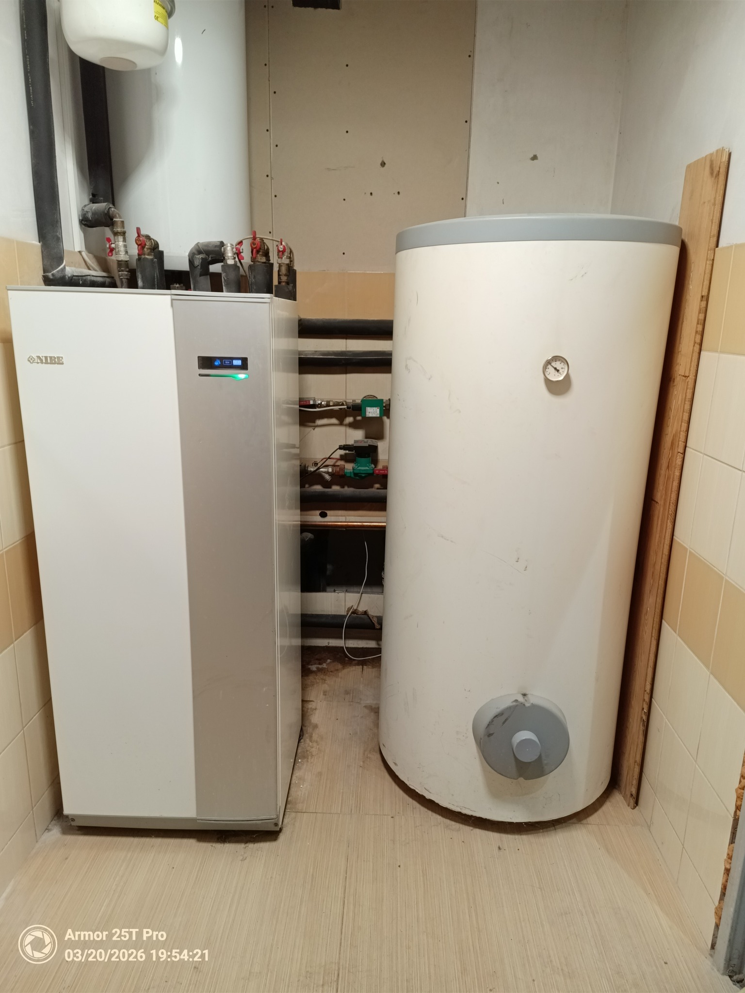 Biała pompa ciepła Nibe i zbiornik akumulacyjny w kotłowni z beżowymi płytkami. Widoczne rury i zawory. Ujęcie z bliska, prezentujące instalację.