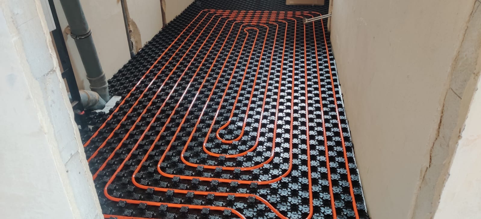 Pomarańczowe rury ogrzewania podłogowego ułożone na czarnej macie w surowym wnętrzu. Widoczne elementy instalacji przy ścianie. Ujęcie z góry, szeroki kadr.