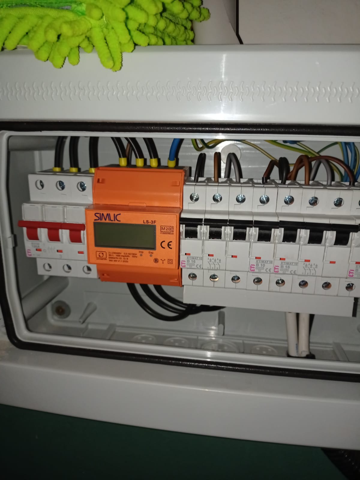 Rozdzielnica elektryczna w obudowie, z widocznymi bezpiecznikami ETIMAT i pomarańczowym modułem SIMLIC LS-3F z wyświetlaczem. Widoczne przewody i elementy montażowe.