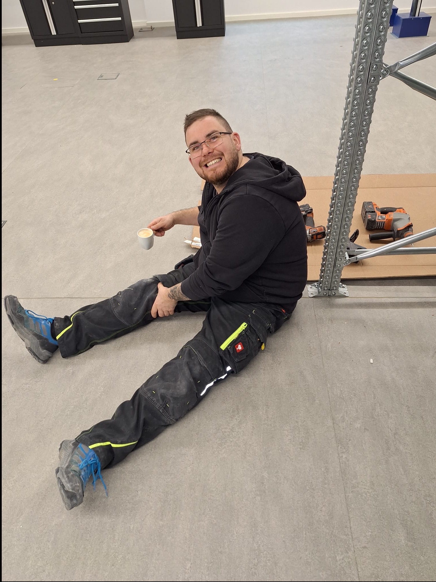 Uśmiechnięty monter w roboczych spodniach, siedząc na podłodze z kawą, obok narzędzi i metalowej konstrukcji regału w jasnym wnętrzu.