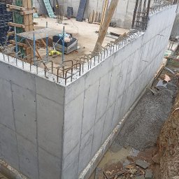 PLASPANEL Ltd - Ściana żelbetowa z wystającymi prętami zbrojeniowymi, widoczna z boku, na placu budowy. W tle rusztowania i materiały budowlane.