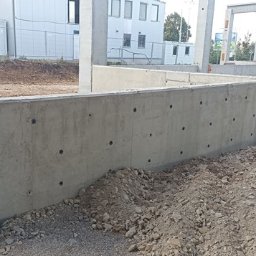 PLASPANEL Ltd - Surowy betonowy fundament z otworami, widok z bliska na placu budowy. W tle budynek z białymi oknami i słupami konstrukcyjnymi.