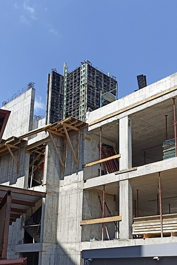 Surowy, żelbetowy szkielet budynku wielopiętrowego w trakcie budowy, z widocznymi deskowaniami i podporami, na tle błękitnego nieba. Ujęcie z dołu.