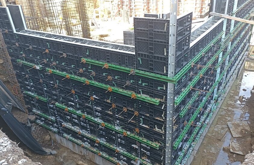 Szczegółowe ujęcie szalunku fundamentowego z tworzywa sztucznego, wzmocnionego zielonymi belkami i metalowymi ściągami, gotowego do zalania betonem.