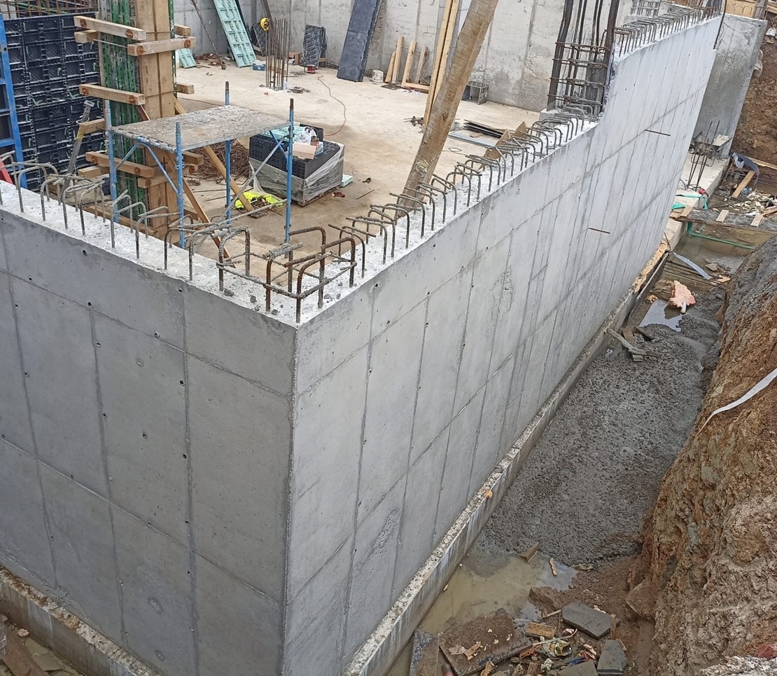 Ściana żelbetowa z wystającymi prętami zbrojeniowymi, widoczna z boku, na placu budowy. W tle rusztowania i materiały budowlane.