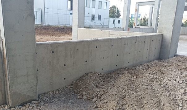 Surowy betonowy fundament z otworami, widok z bliska na placu budowy. W tle budynek z białymi oknami i słupami konstrukcyjnymi.