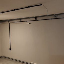Krzysztof Wilk Elektryka i Automatyka Domowa - Szara ściana z widoczną instalacją elektryczną w czarnych rurkach, poprowadzona po suficie i ścianach. Widoczny fragment grzejnika i podłogi z szarych płytek.