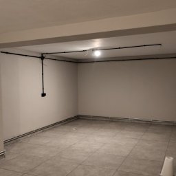 Krzysztof Wilk Elektryka i Automatyka Domowa - Wykończone wnętrze z widoczną, natynkową instalacją elektryczną w czarnych rurkach. Na ścianach gniazdka i włączniki. Pod sufitem lampa. W tle widoczne worki z zaprawą.